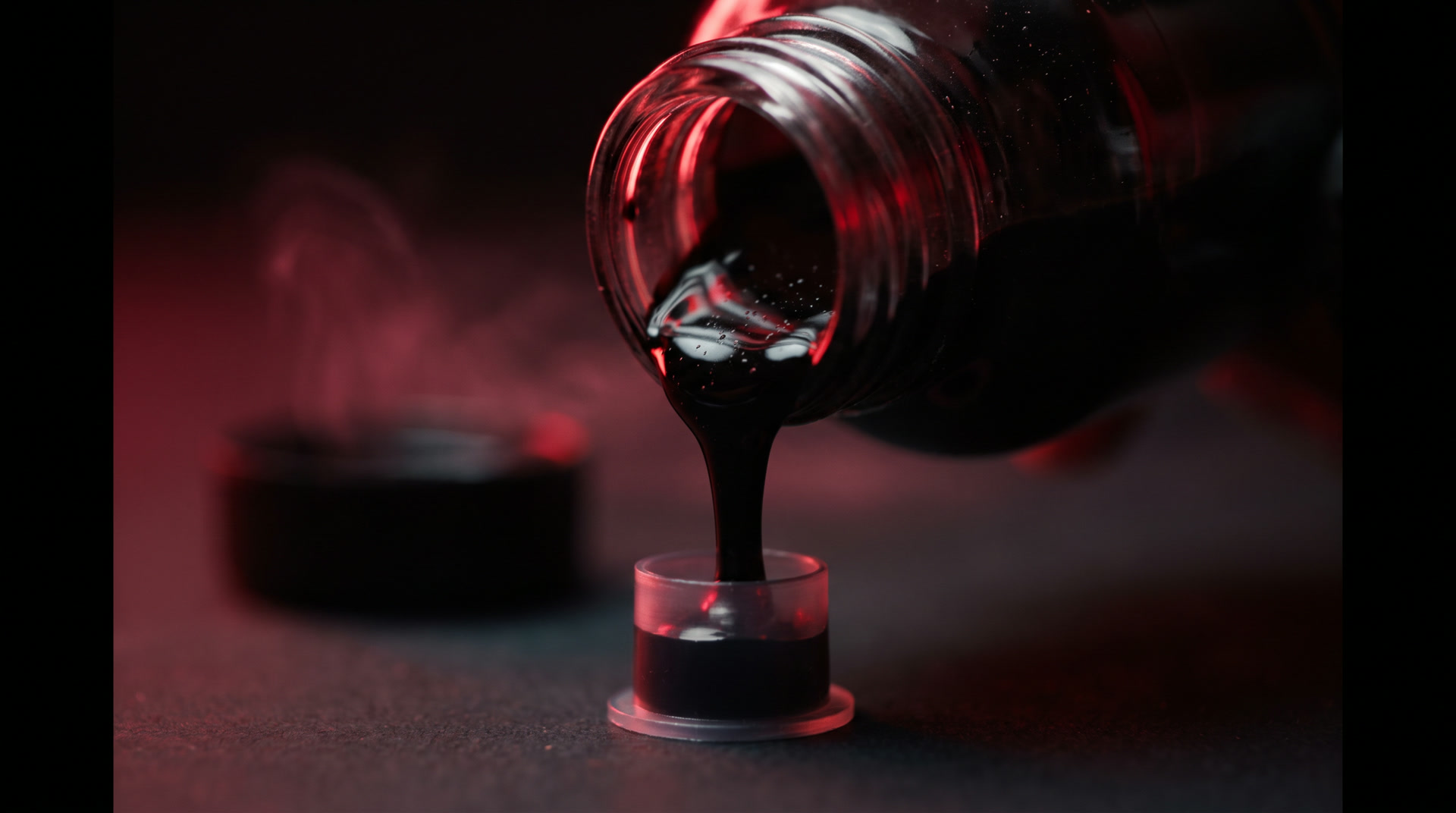 Black tattoo ink pouring from a bottle into an ink cap under red light